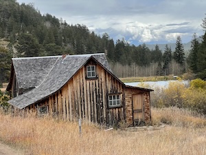 A rustic cabin sits in front of a small mountain lake with pine trees in the background.