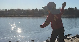 A young child throws a rock into a lake as the sun reflects off the water.